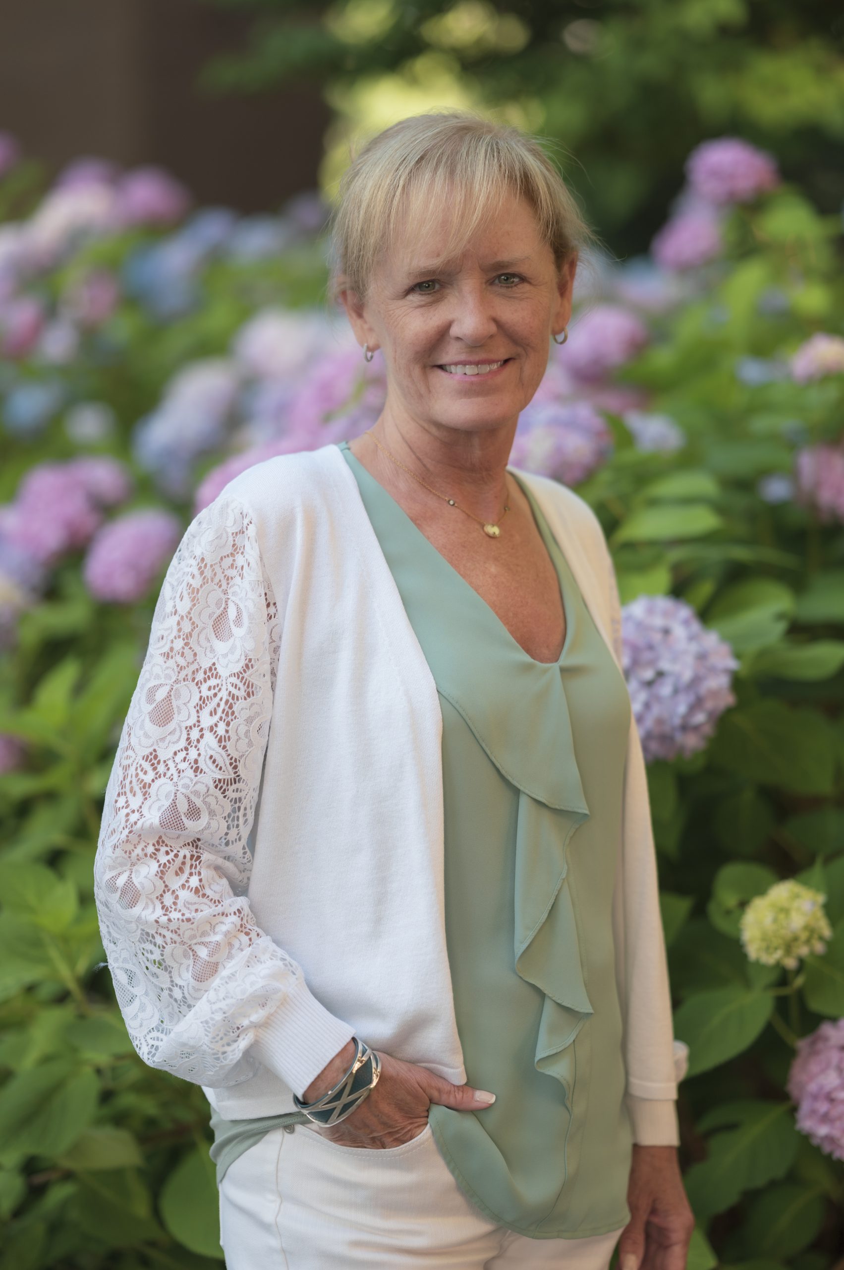 A woman smiles while standing amidst colorful hydrangea flowers. She wears a light green blouse and a white cardigan with lace details, exuding a warm, inviting presence.
